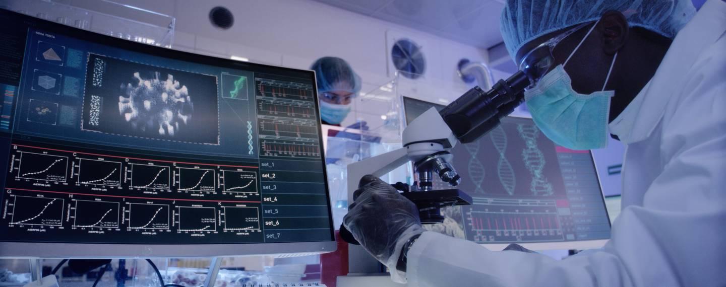 A researcher looks into a microscope next to a computer displaying a covid cell