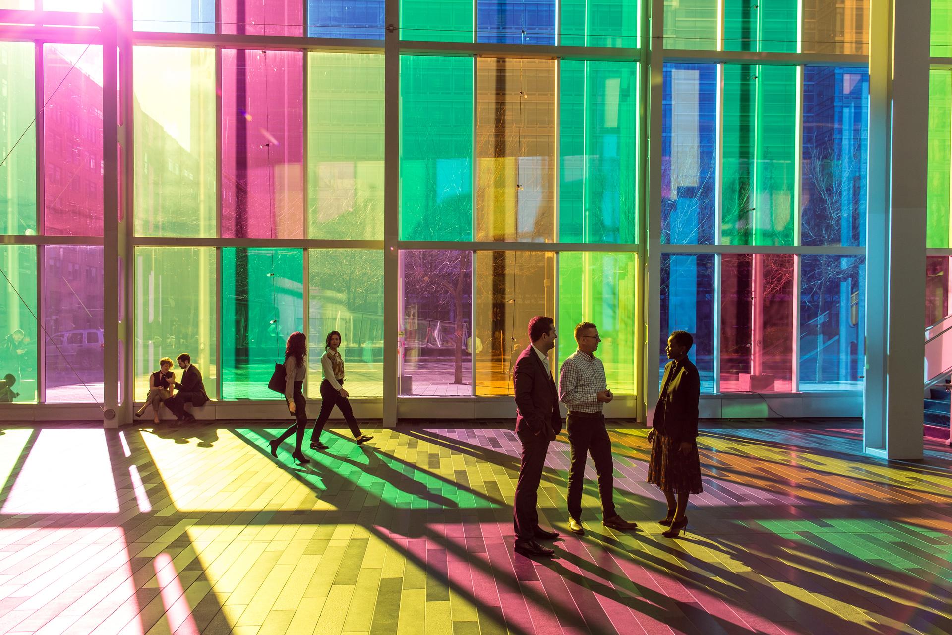 Groups of people in a large hallway of colourful reflective glass windows.
