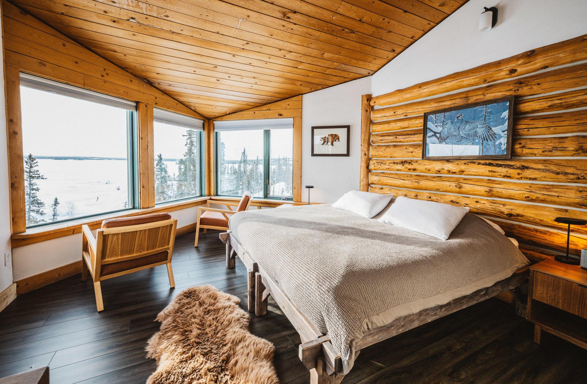 Cozy log cabin bedroom with large windows overlooking a snowy lake and forest landscape