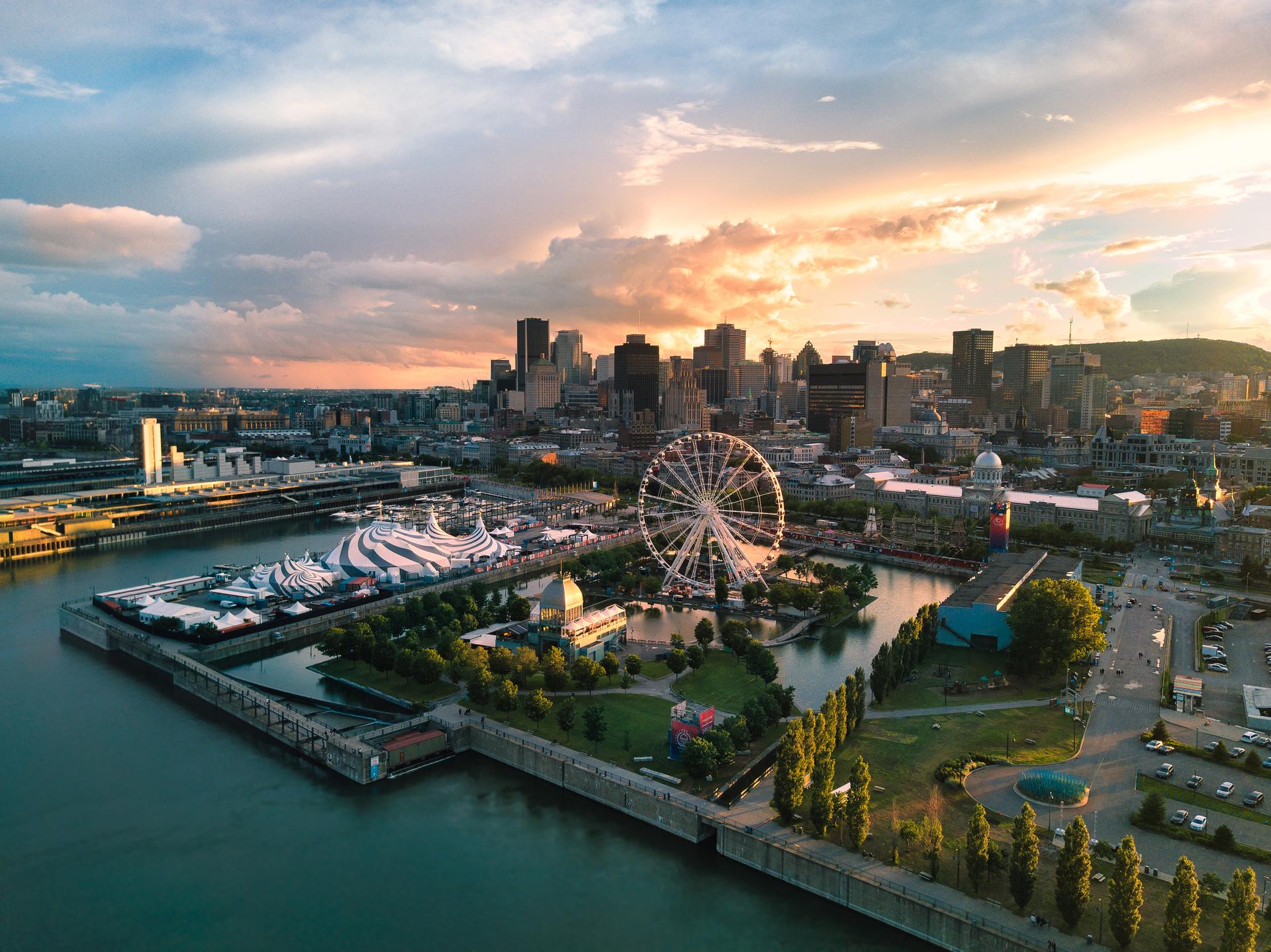 Aerial sunset view of Old Montreal's waterfront along the St. Lawrence River, featuring the Montreal Observation Wheel, Bonsecours Market dome, festival tents, and the downtown skyline with Mont Royal in the background.